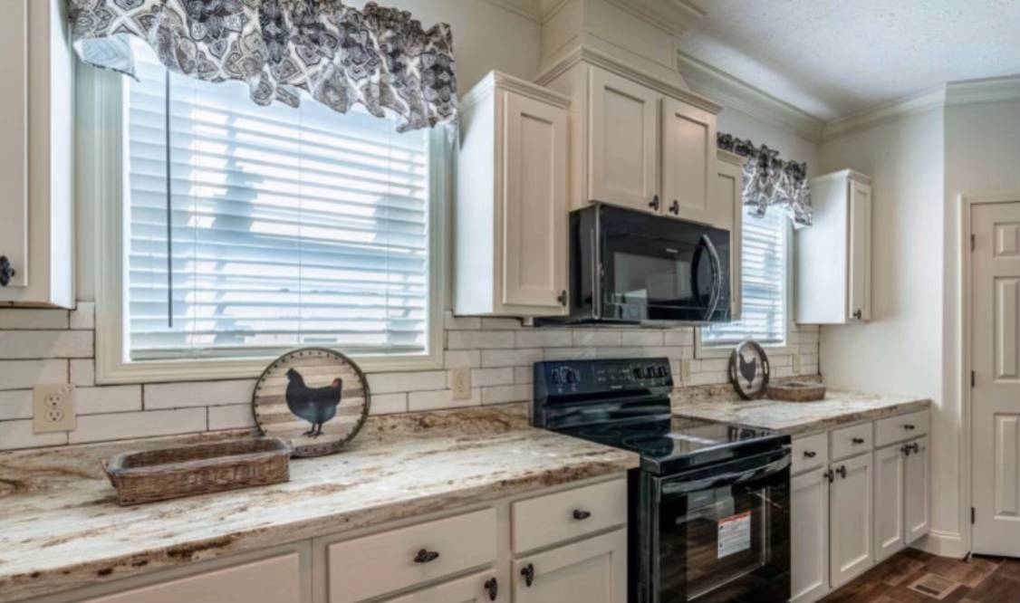 Modern kitchen with white cabinets, black appliances, and patterned curtains on windows. The countertop features a rustic tray and decorative plates. The overall tone is bright and inviting.