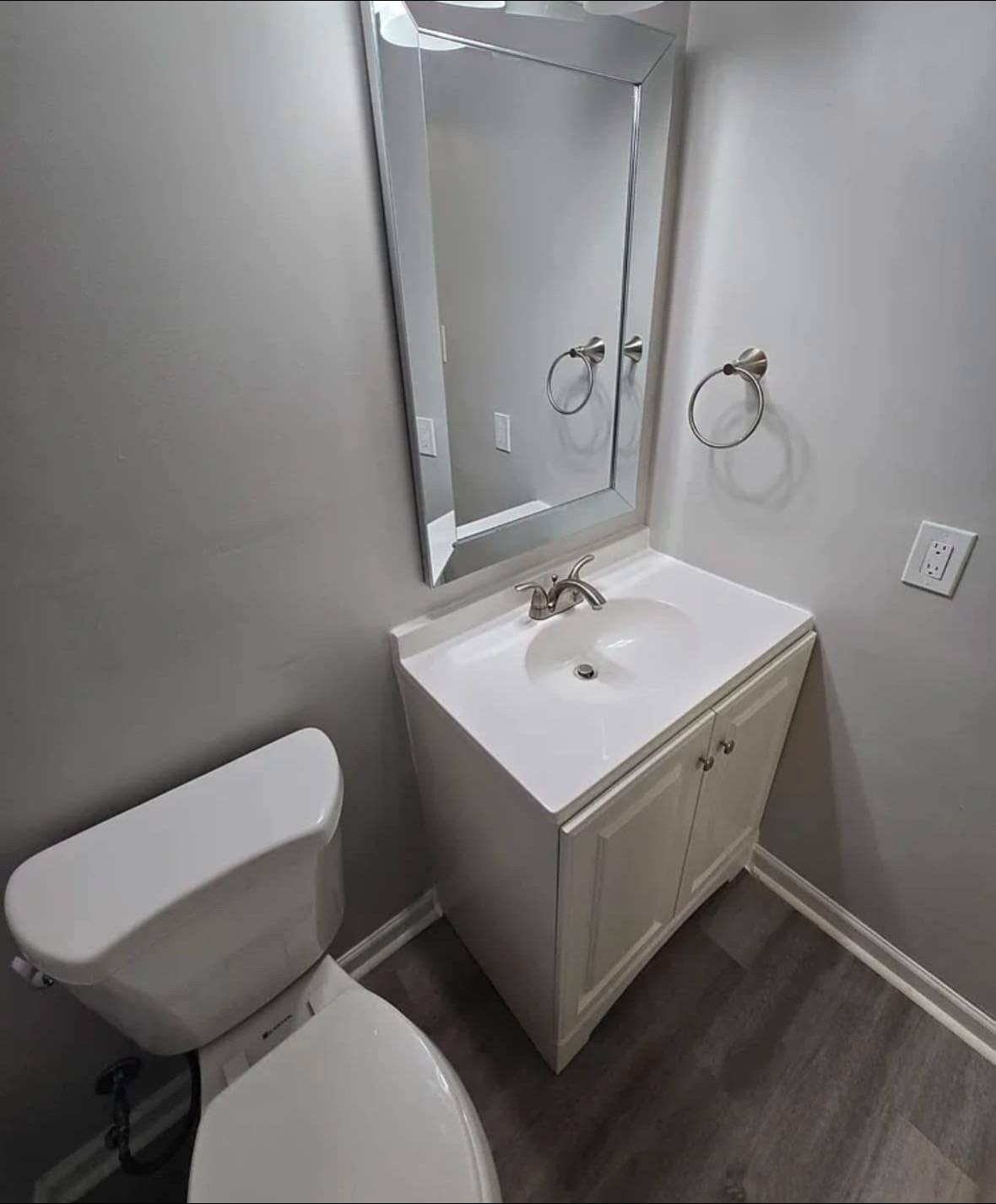 Compact, modern bathroom with a white toilet and sink vanity, accented by a silver mirror and towel ring, set against gray walls and wood floor.