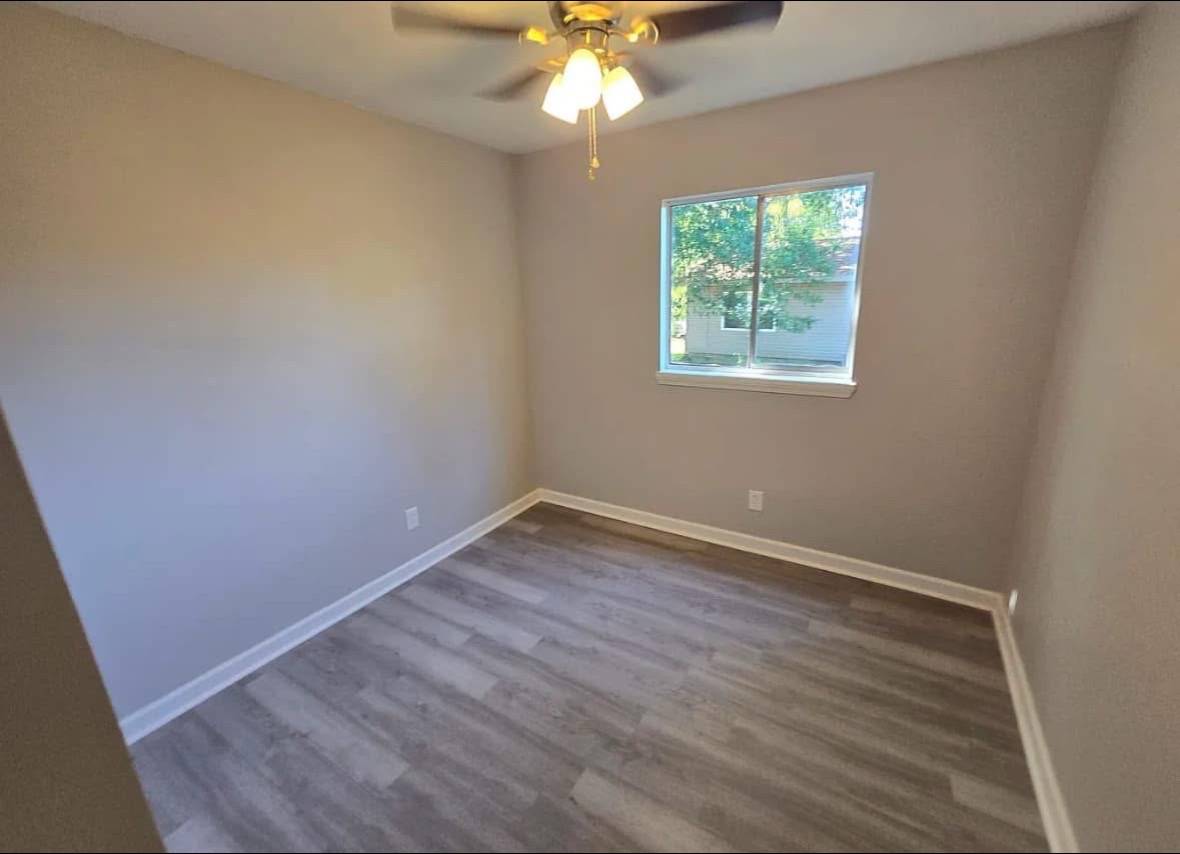 Small, empty room with light gray walls and wood flooring, featuring a ceiling fan with lights. A window offers a view of leafy greenery outside.