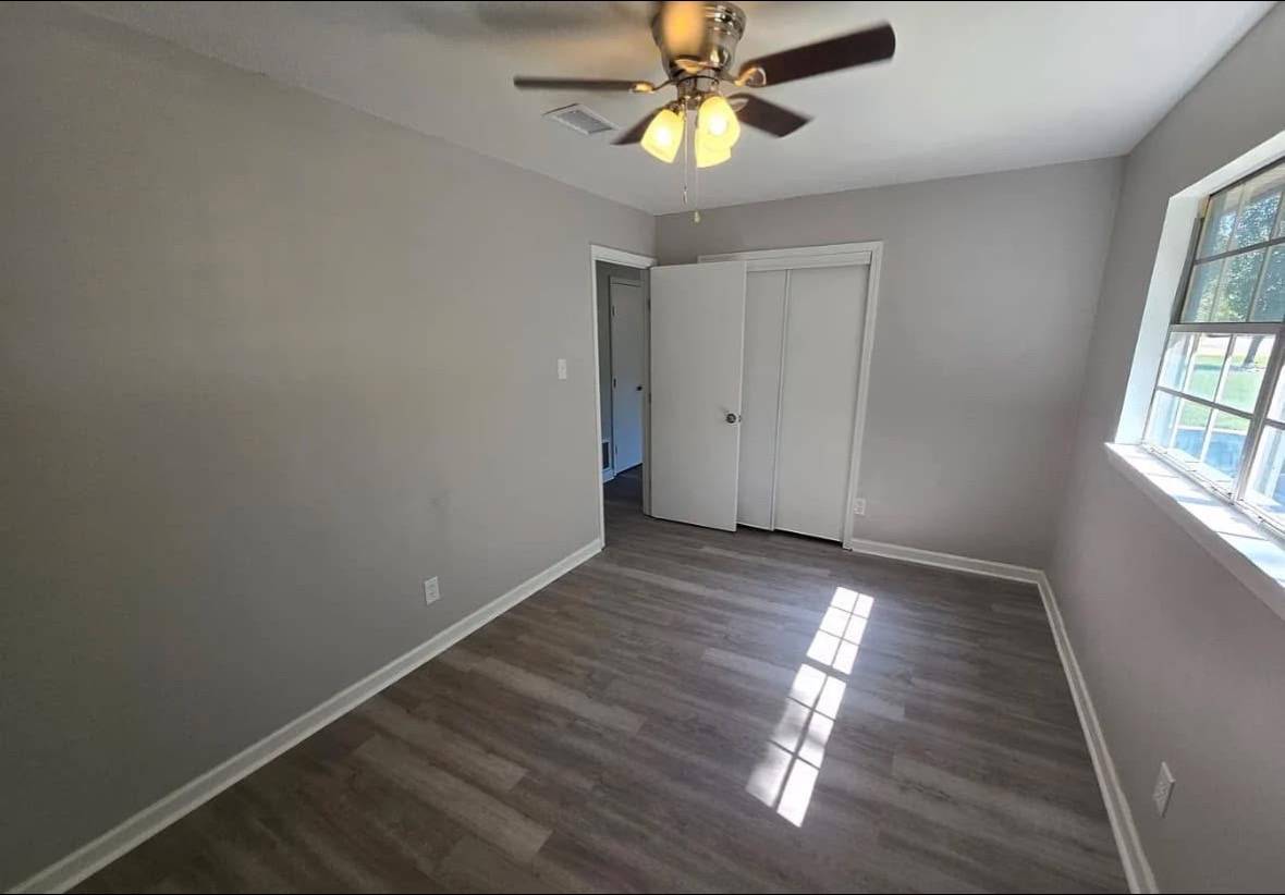 Small empty room with gray walls and wood laminate flooring. A ceiling fan with lights is above, and natural light streams through a window on the right.
