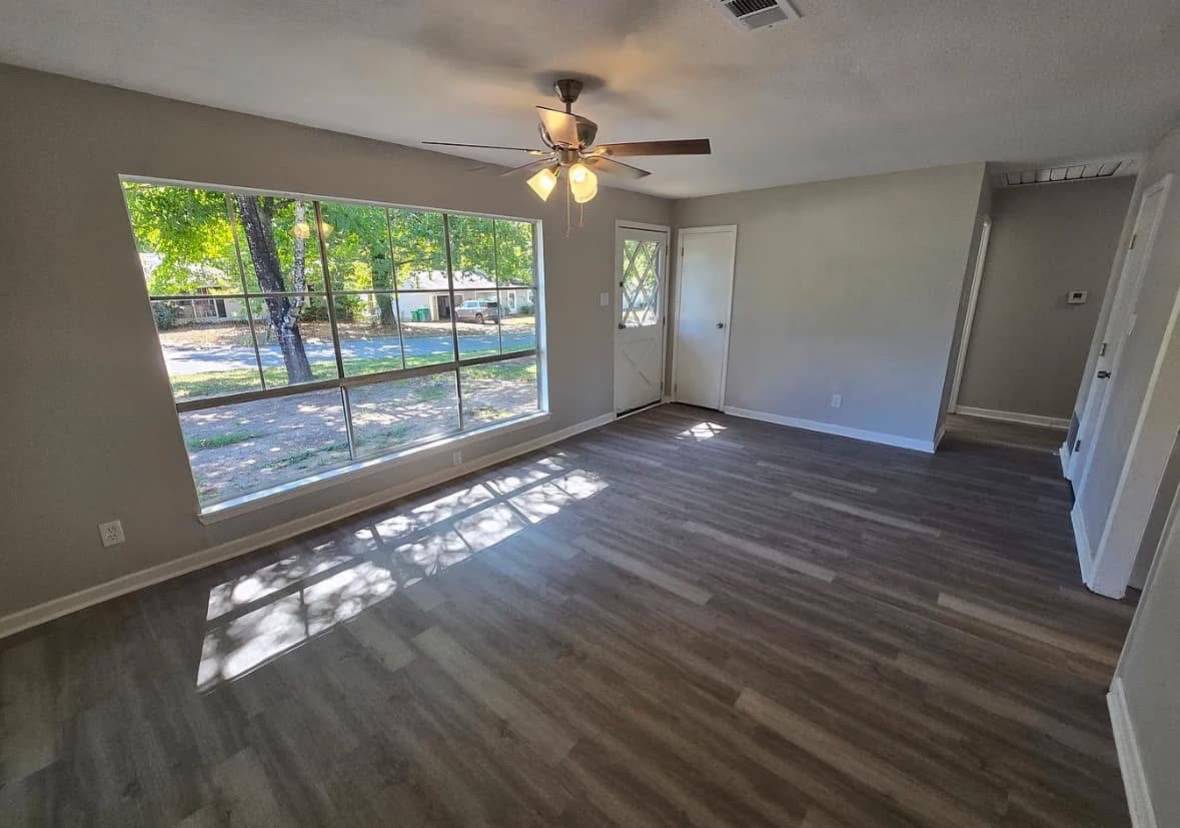 Spacious living room with light gray walls and wood flooring. Large window with a view of green trees. Ceiling fan and a door leading outside. Bright, airy atmosphere.