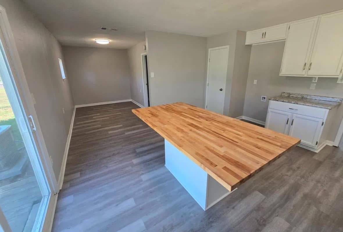 Modern kitchen with light gray walls and wood flooring, featuring a large wooden countertop island. White cabinets are mounted above a granite counter.