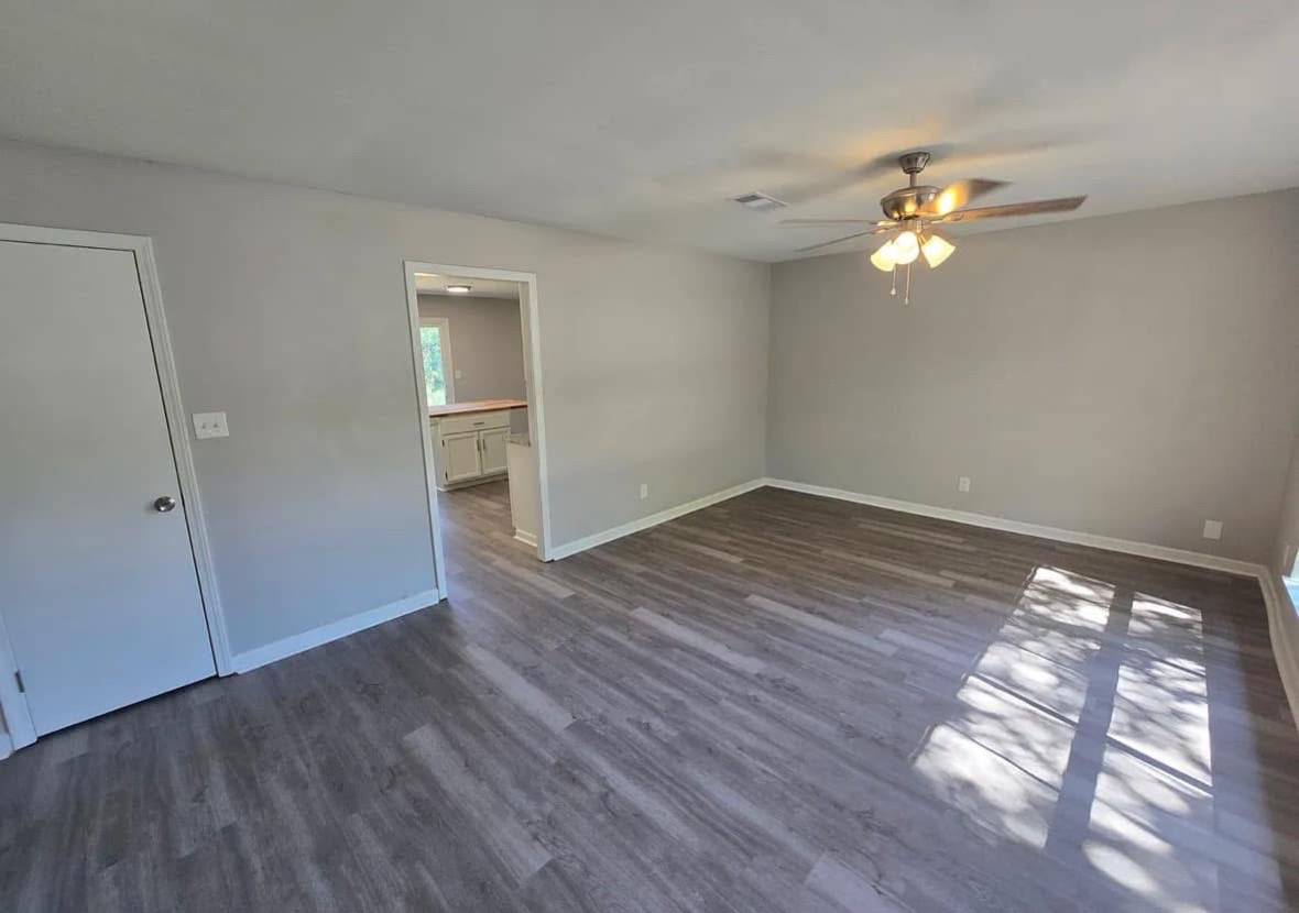 Bright room with gray laminate floors, a ceiling fan, and neutral walls. Sunlight streams in, creating a warm, inviting atmosphere. A doorway leads to a kitchen.
