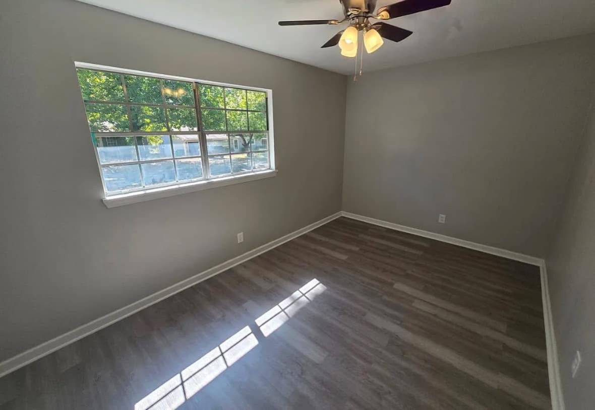 A small, empty room with gray walls and wood flooring is lit by natural light from a window showing green trees. A ceiling fan with lights hangs above.