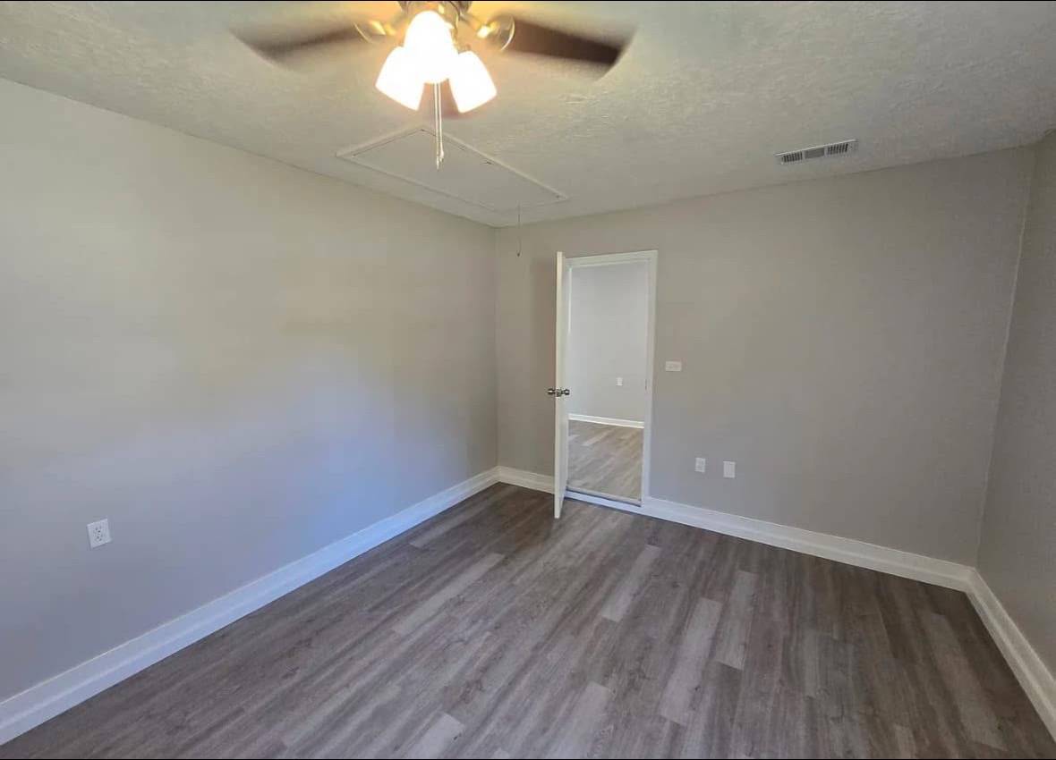A vacant room with light gray walls, light brown wood flooring, and a ceiling fan with lights. An open door leads to another empty room. Calm, simple atmosphere.