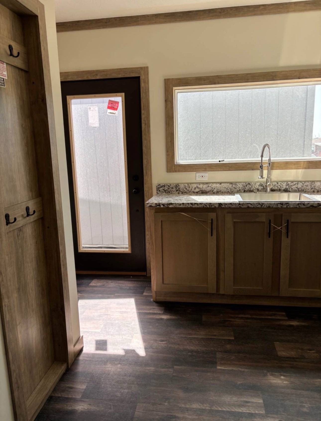 A kitchen corner with wood cabinets and granite countertop under a large window. A glass-paneled door lets in sunlight, highlighting the dark wood floor.