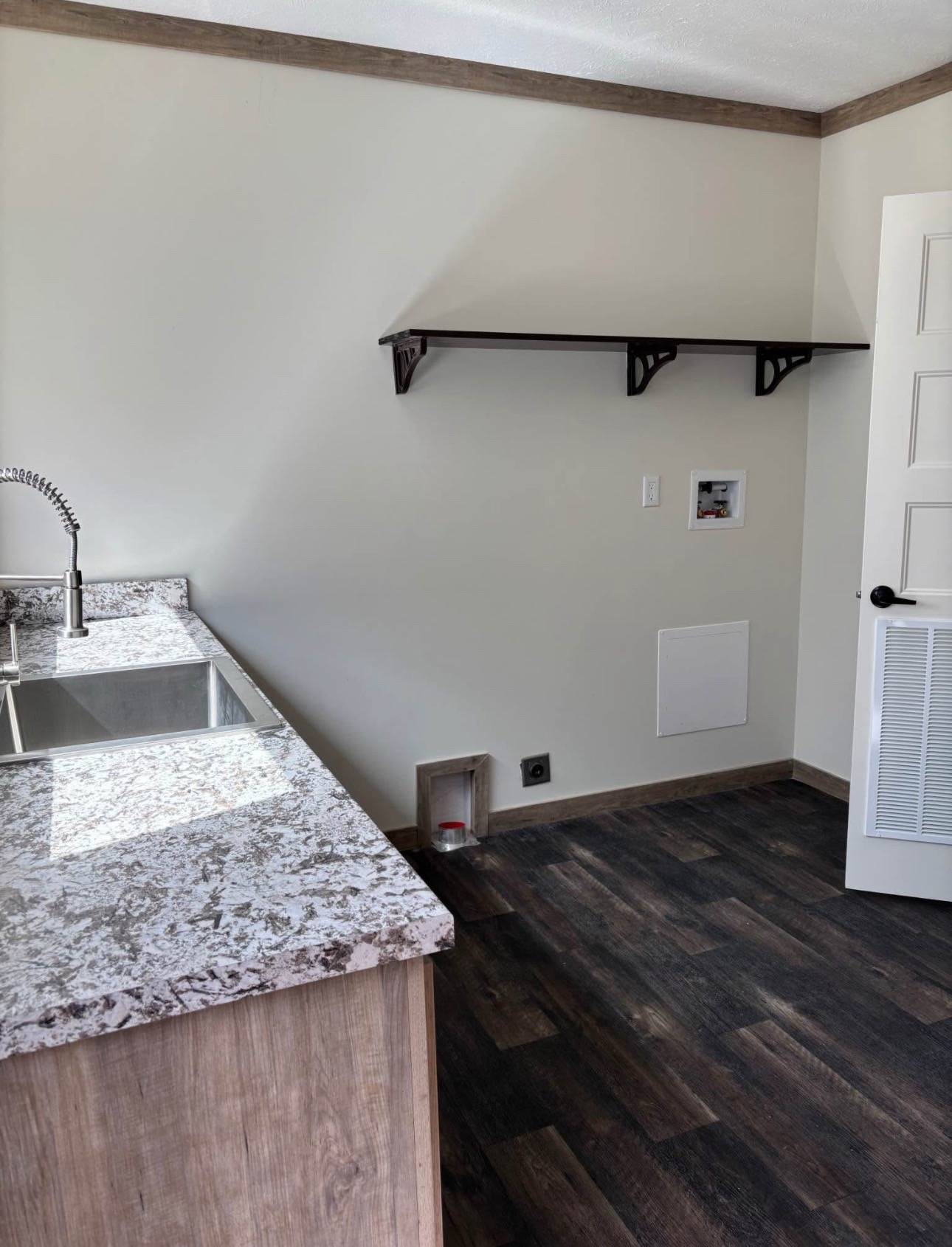 A modern laundry room with dark wood flooring, a marble countertop sink, and a black shelf on a light beige wall. The atmosphere is clean and minimalist.