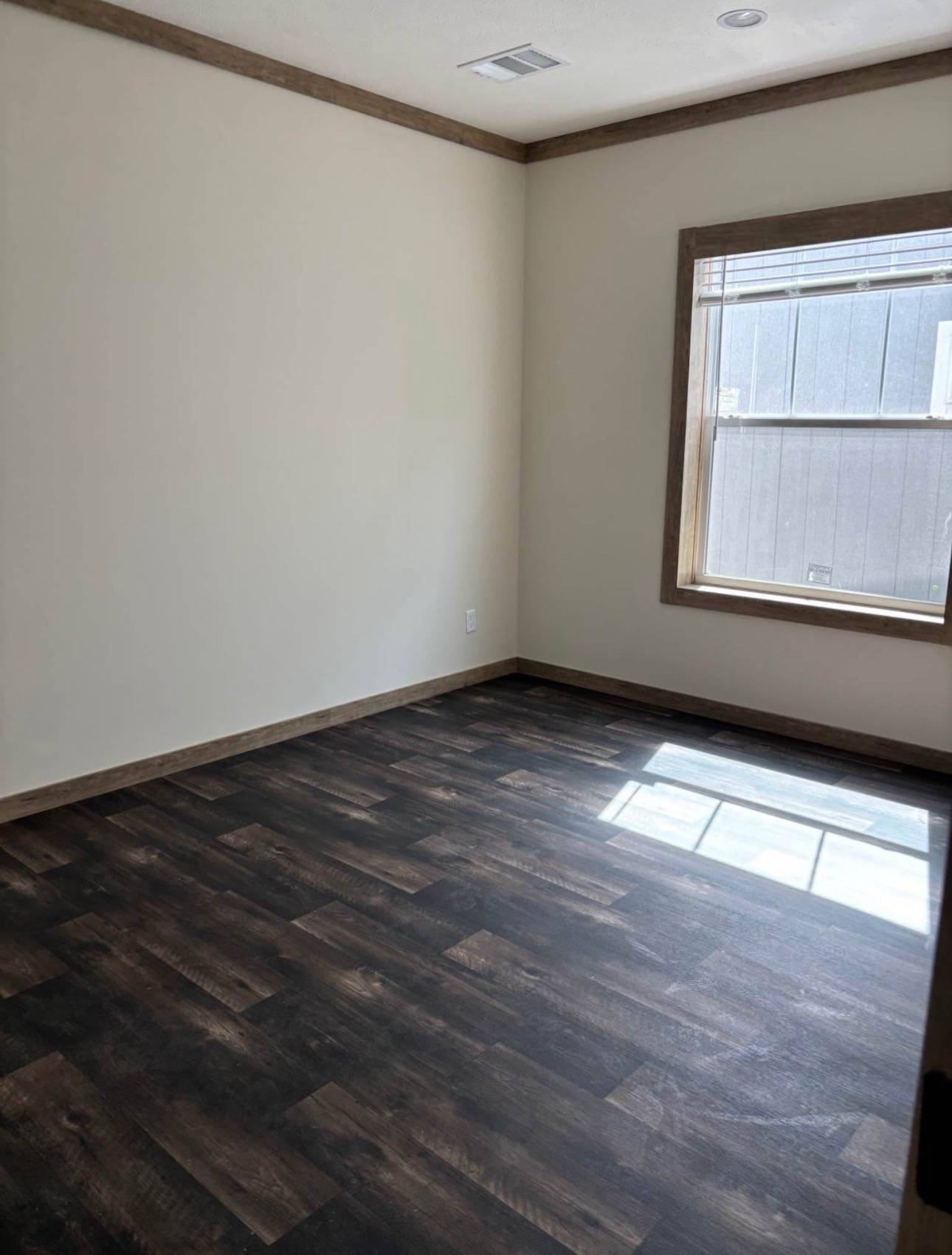 An empty room with beige walls and dark wood flooring. A single window on the right lets in natural light, casting a soft shadow on the floor.