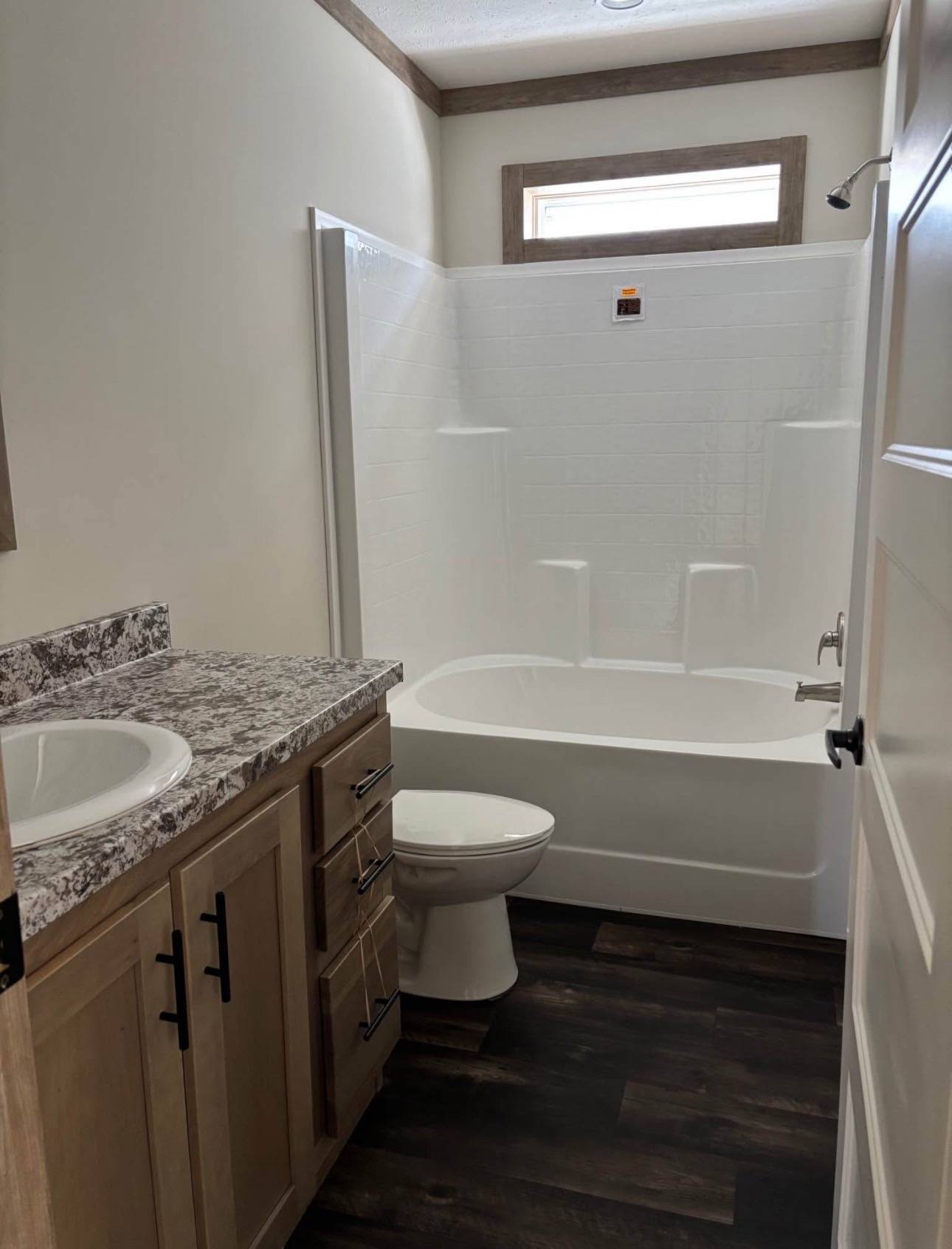 A small bathroom with a white bathtub and shower, granite countertop with sink, wooden cabinets, dark floor, and natural light from a window above.
