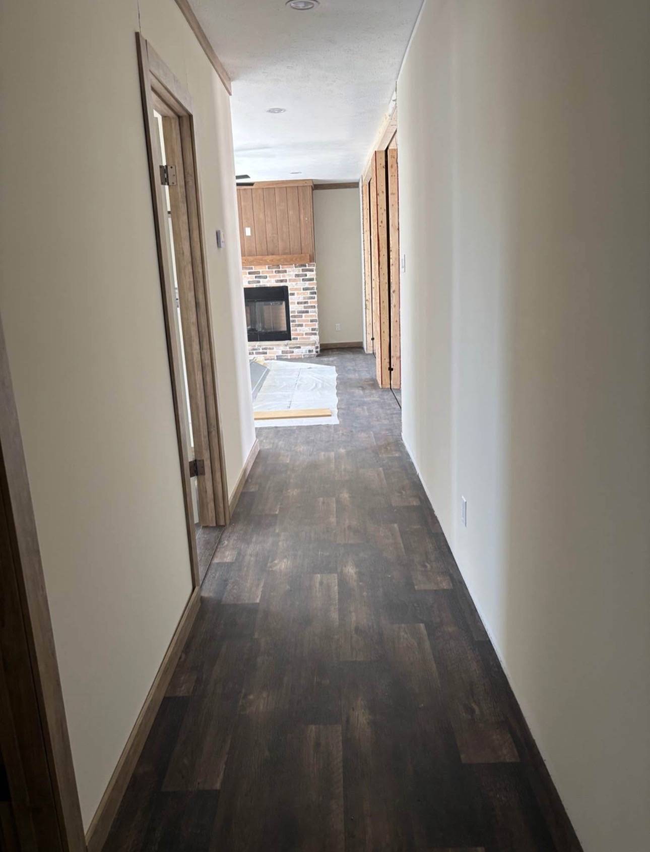 A narrow hallway with dark wood flooring leads to a brick fireplace in a cozy room. The walls are painted light beige, and a wooden door is partially open.