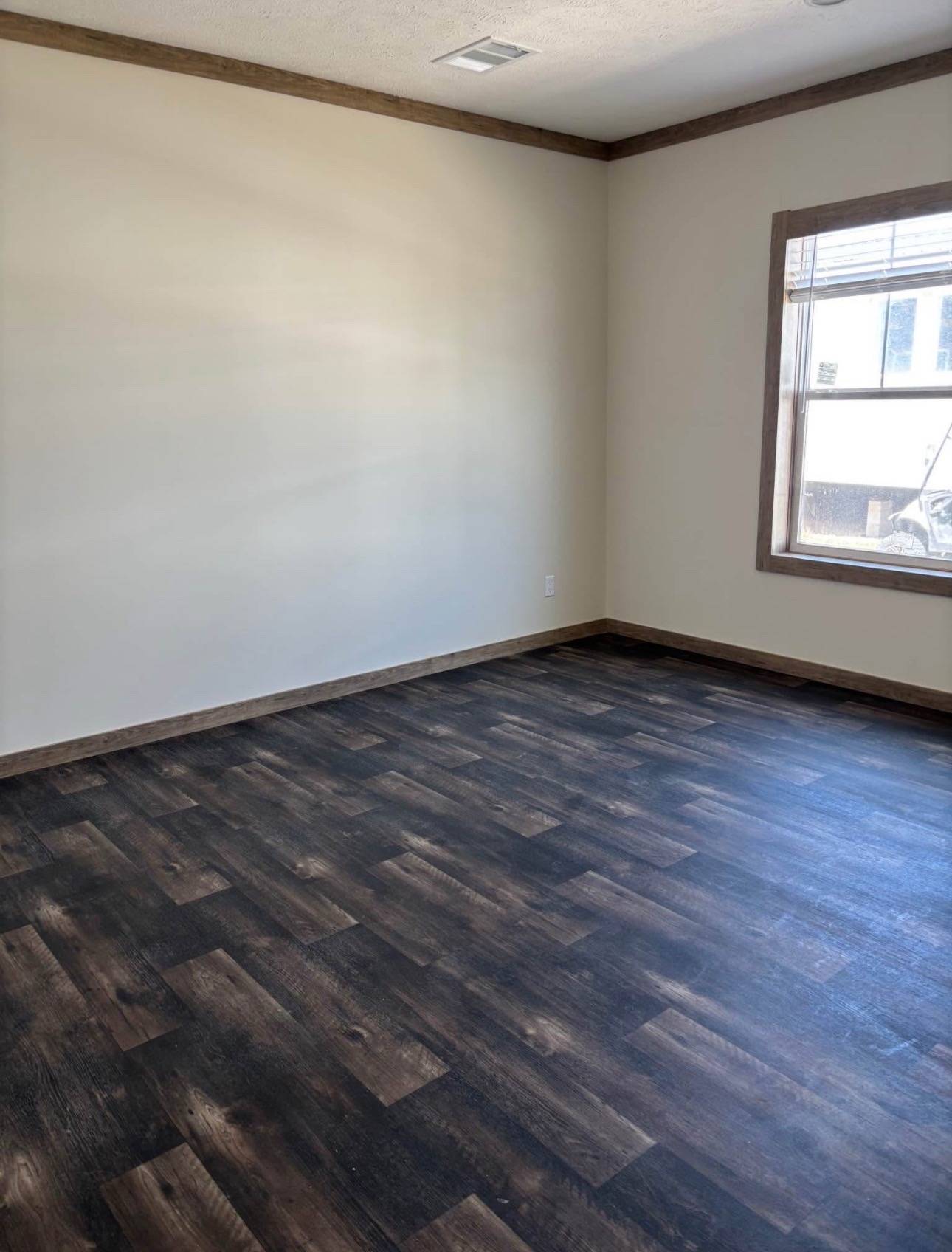 Empty room with dark wood laminate flooring, beige walls, and a window on the right letting in natural light. The space feels clean and unused.