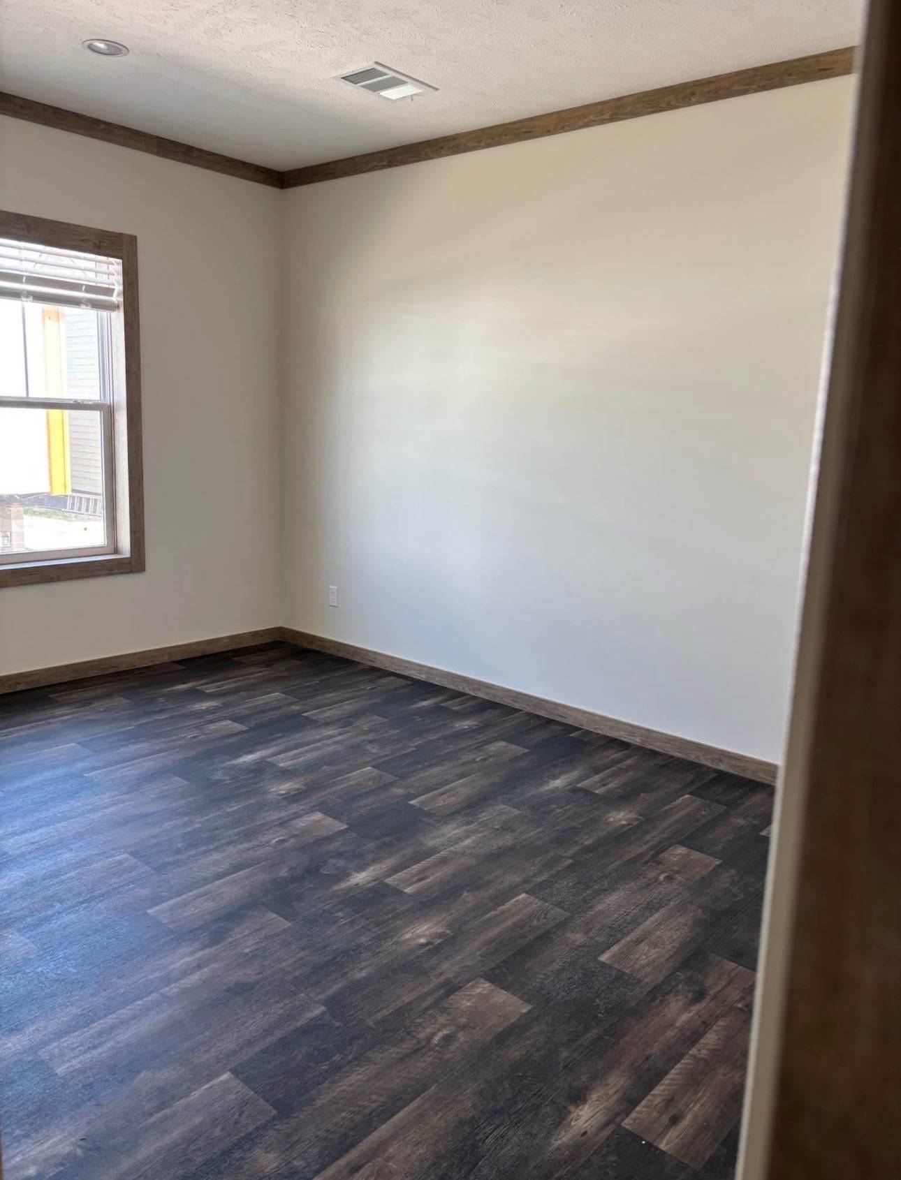 Empty room with dark wood flooring, beige walls, and a window with wooden trim. Natural light enters, creating a calm and neutral tone.