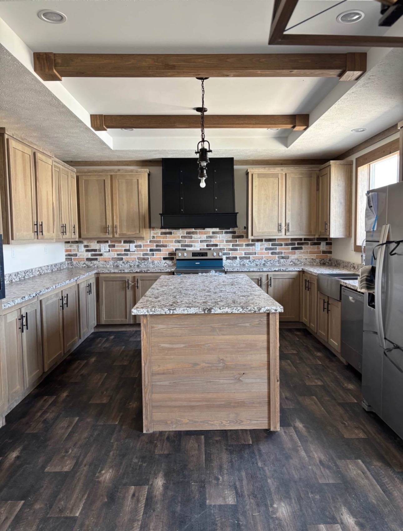 Spacious kitchen with wooden cabinets and beams, a marble island, dark wood floor, and a brick backsplash. Stainless steel appliances add a modern touch.