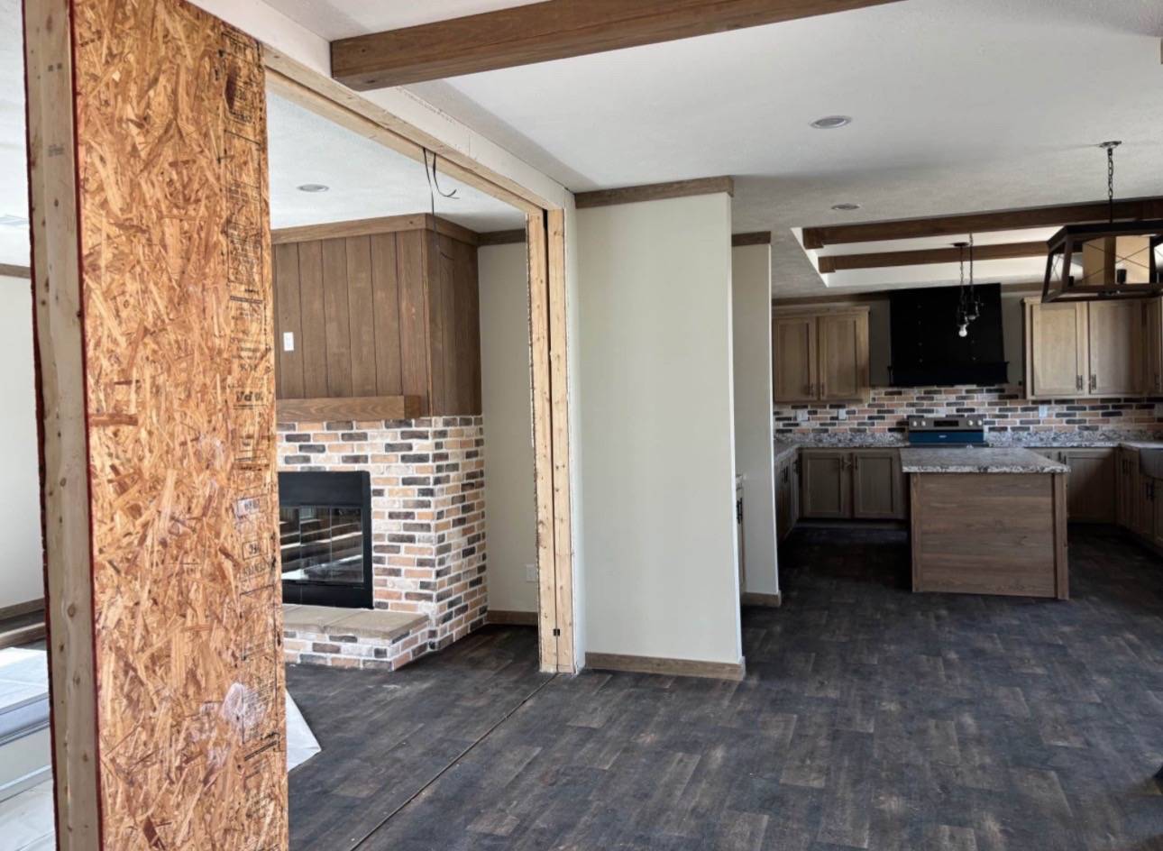 Open-concept kitchen and living area under renovation. Exposed wooden beams and brick fireplace with beige walls. Warm, unfinished, rustic tone.