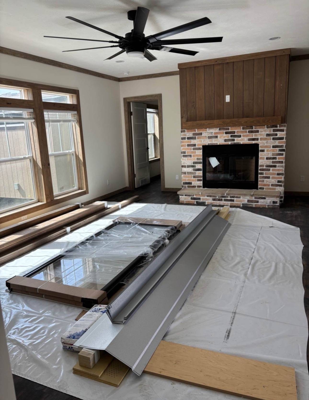 A room under renovation with a large ceiling fan and a brick fireplace. Windows let in natural light. Construction materials are on a white covering.