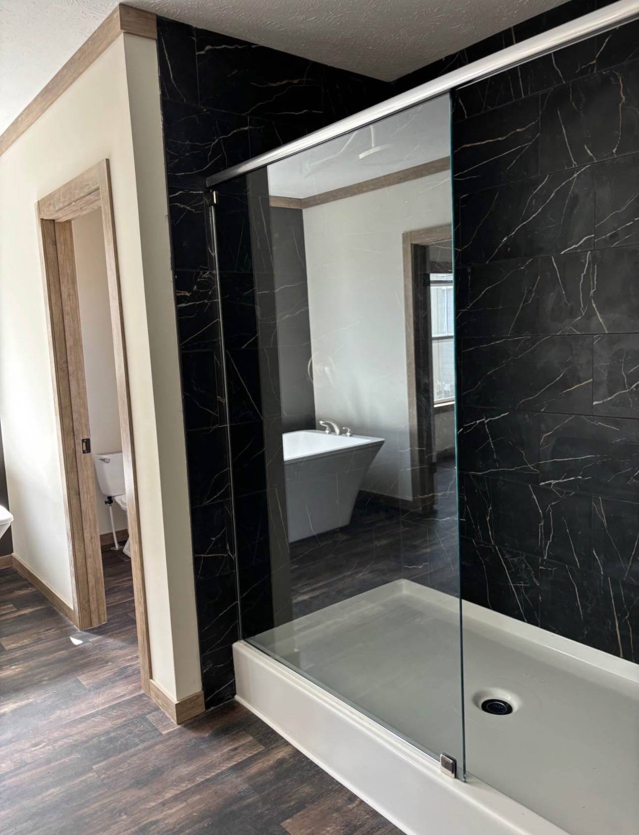 Modern bathroom with dark marble walls, a glass shower enclosure, and a white standalone tub. Wood flooring and trim add warmth. Bright, serene atmosphere.