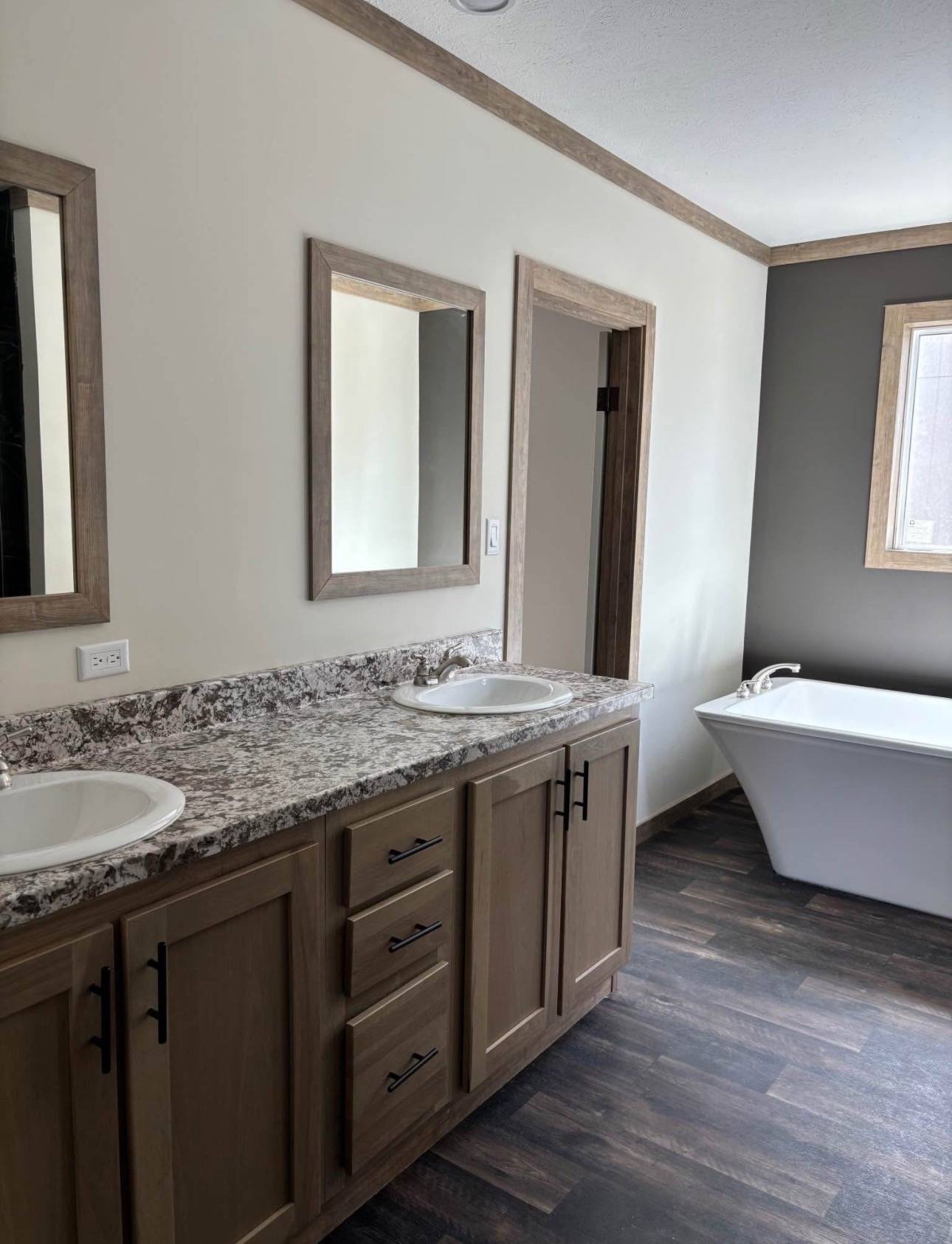 Modern bathroom with dark marble walls, a glass shower enclosure, and a white standalone tub. Wood flooring and trim add warmth. Bright, serene atmosphere.