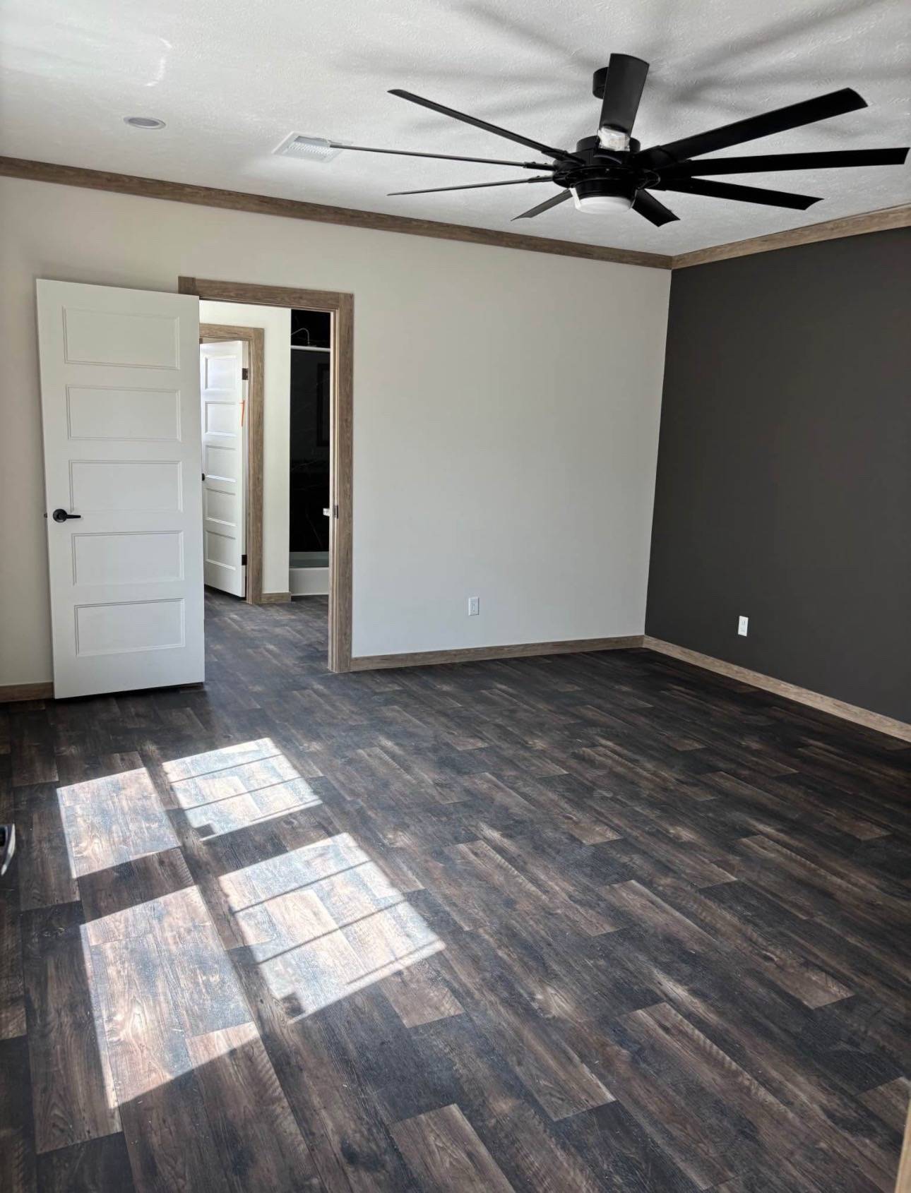 A room with dark wood flooring and a large black ceiling fan. The white door is slightly open, leading to a hallway. Sunlight casts patterns on the floor, creating a calm and inviting atmosphere.
