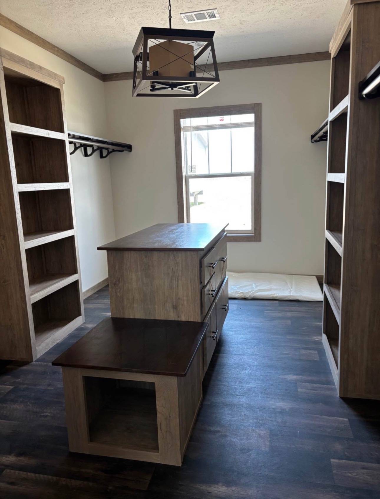 Spacious walk-in closet with wooden shelves and a central island featuring drawers and a bench. It has dark flooring and a large window, creating a modern, airy atmosphere.