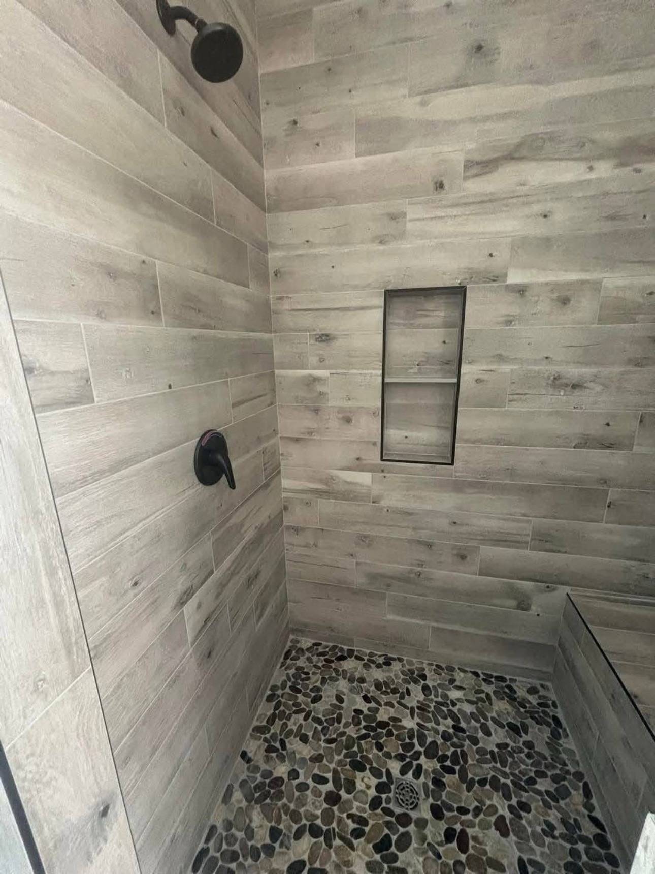 Walk-in shower with gray wooden-style tile walls, black shower fixtures, and a pebble stone floor. Built-in rectangular shelf adds a modern touch.