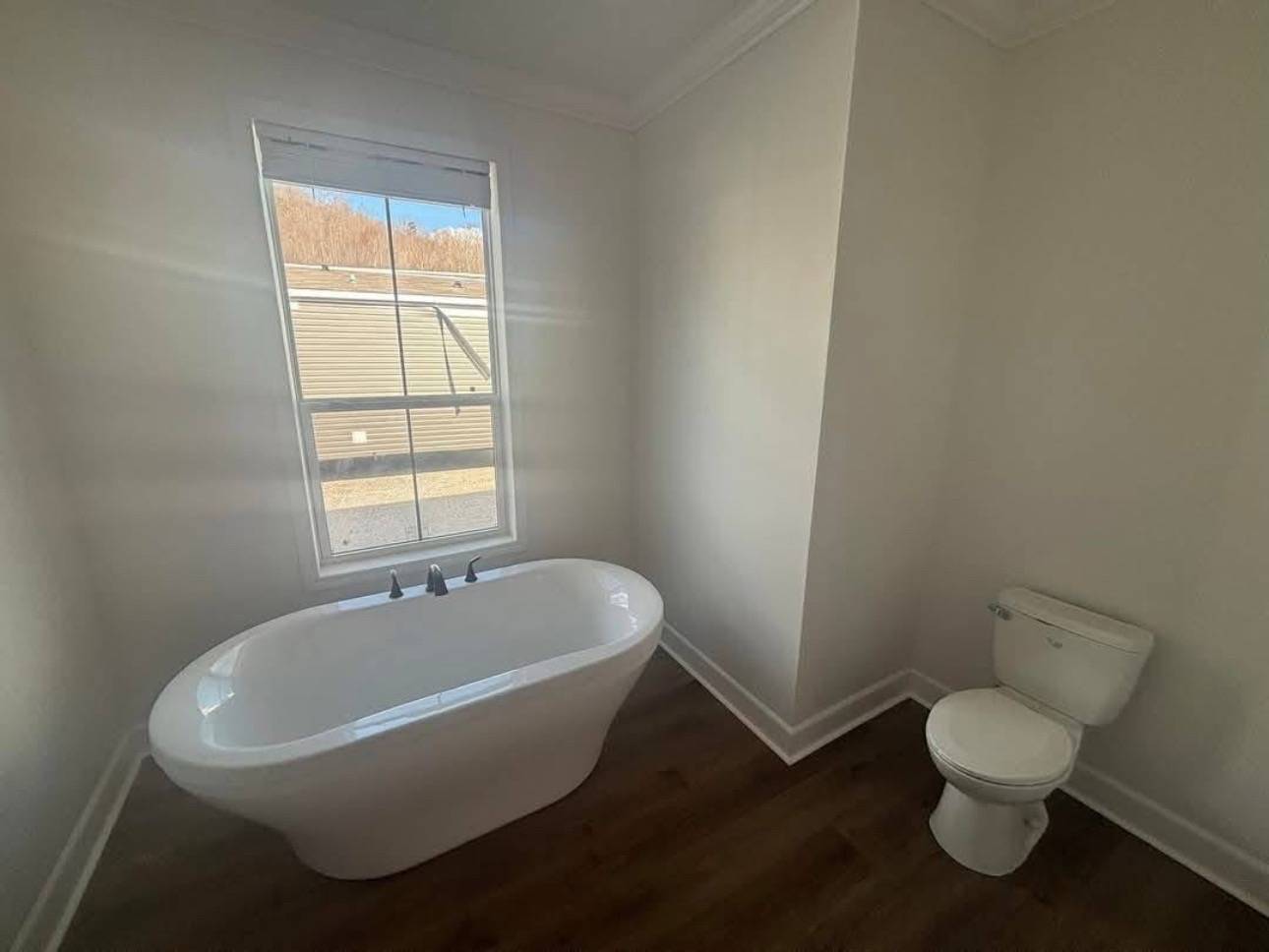 Minimalist bathroom with a white freestanding bathtub under a window and a toilet to the side. The room is bright with natural light and has wooden flooring.