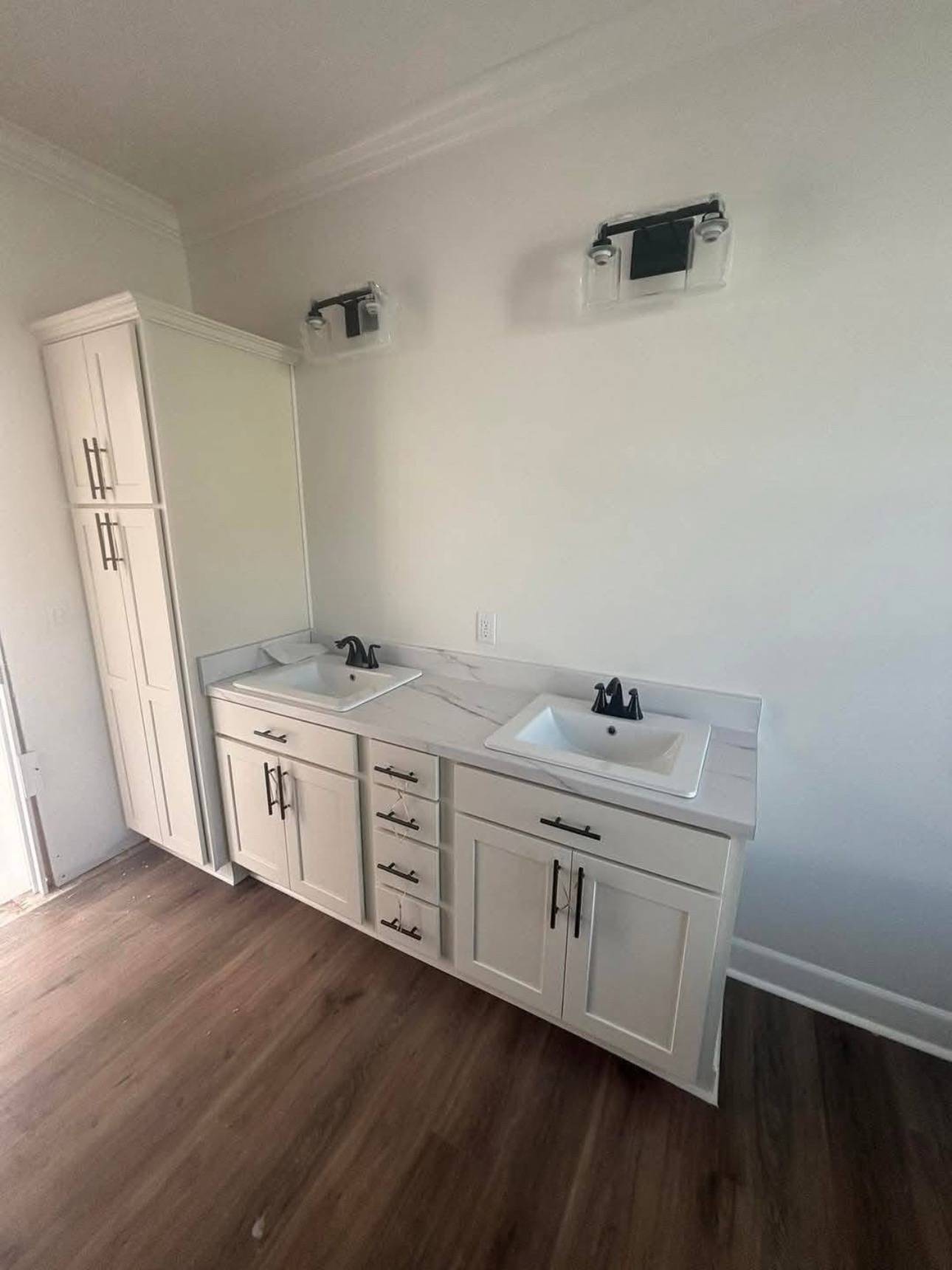 Modern bathroom with a dual-sink white vanity, black faucets, and elegant marble countertop. Tall cabinet on the left; wood flooring adds warmth.