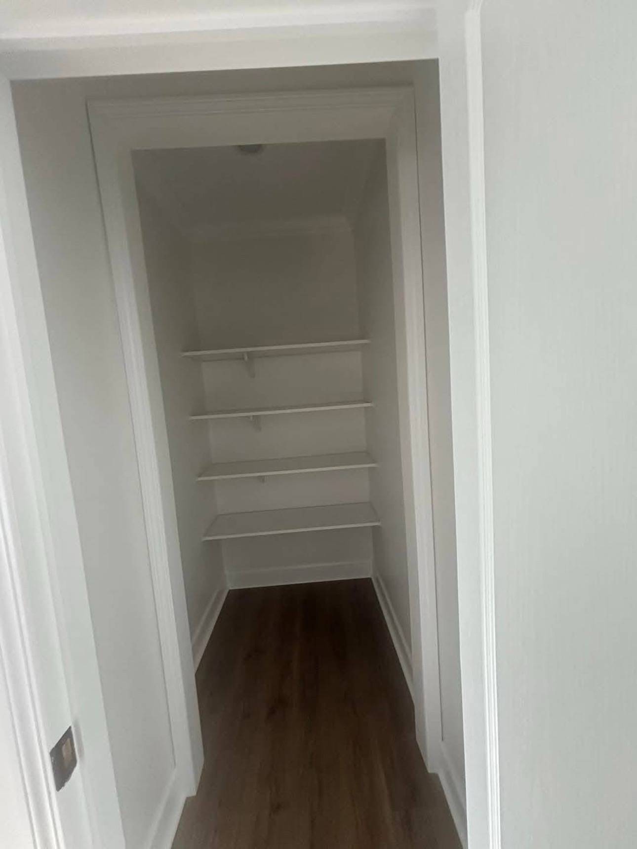 A small empty closet with white walls and three narrow shelves. The floor is wooden, adding a warm contrast to the otherwise minimalist space.