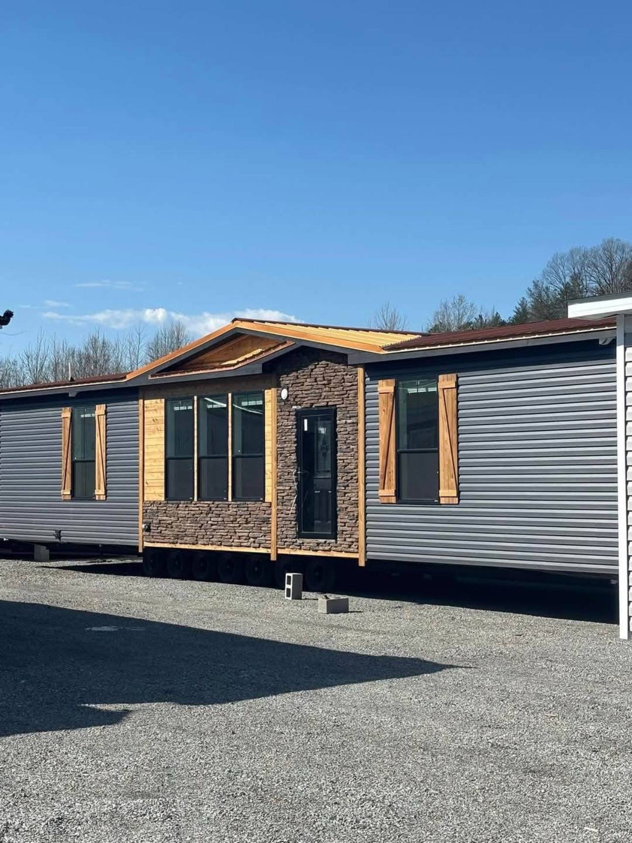 A modern modular home with gray siding and stone accents sits on a gravel lot. Wooden shutters and a black door add rustic charm under a clear blue sky.
