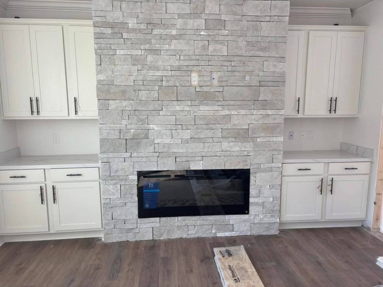 Stone fireplace wall with a modern electric insert, flanked by white cabinets and countertops. Flooring is light wood, creating a cozy, minimalist feel.