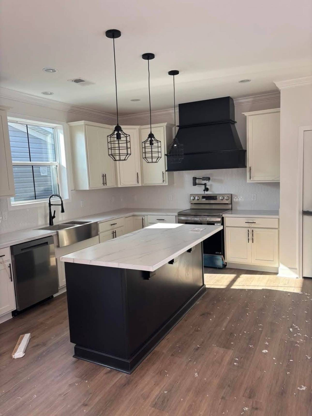 Modern kitchen with white cabinets, black island with marble top, and three pendant lights. Stainless appliances and a black range hood add contrast.