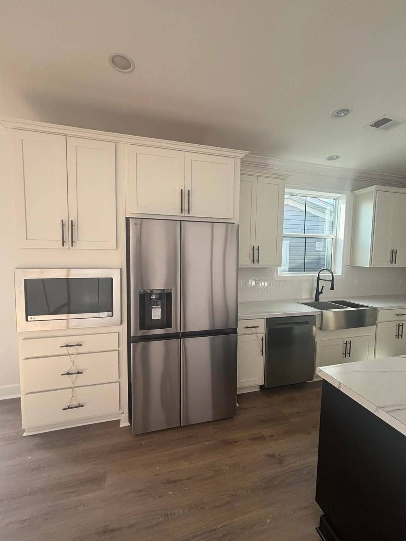 Modern kitchen with stainless steel fridge, built-in microwave, and dishwasher. White cabinets, black handles, and a farmhouse sink under a window.