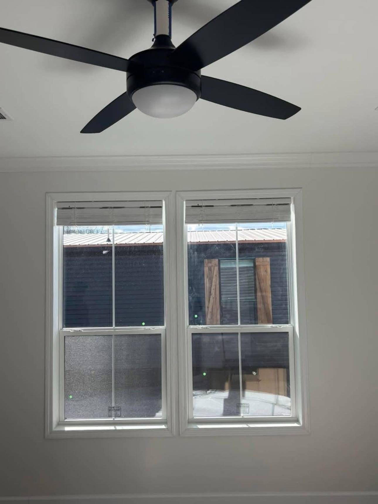 Ceiling fan with black blades above two windows showing an outdoor view of a dark building with wooden shutters, creating a calm interior ambiance.