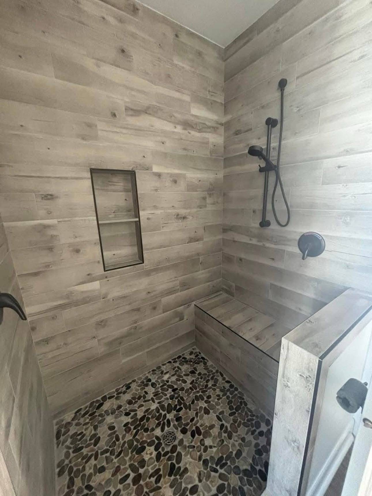 Bathroom shower with textured wood-style walls, black fixtures, and pebble stone floor. Built-in shelf and corner bench add functionality.
