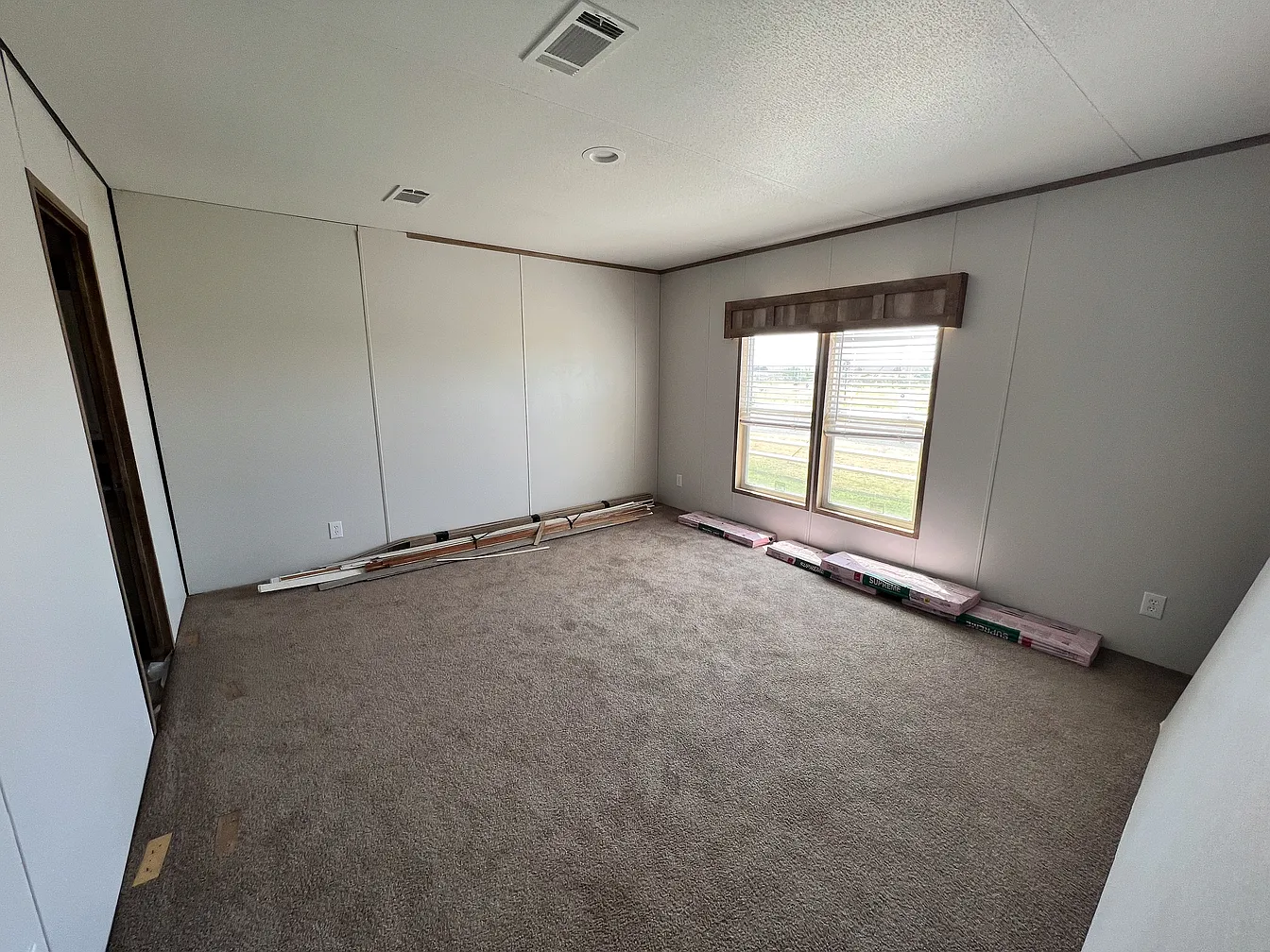 Empty room with beige carpet, light gray walls, and a small window covered by a wooden valance. Flooring materials are stacked along the wall.