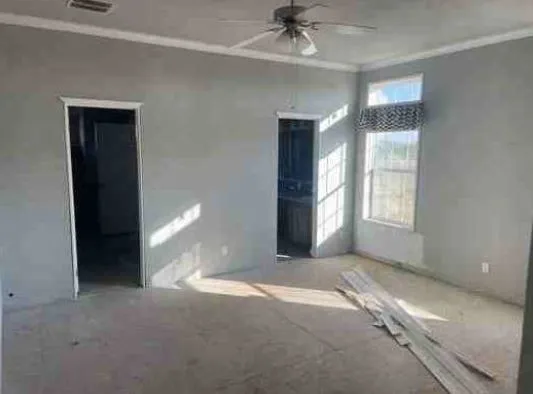 Empty room with gray walls and two doorways. Sunlight streams through a window with blinds onto the carpet. Ceiling fan overhead, wood planks on the floor.