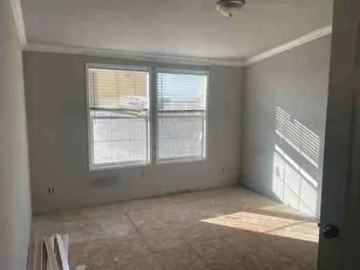 A small, empty room with unpainted walls and bare floors. Two large windows let in sunlight, creating shadows on the floor. Peaceful and minimalistic.