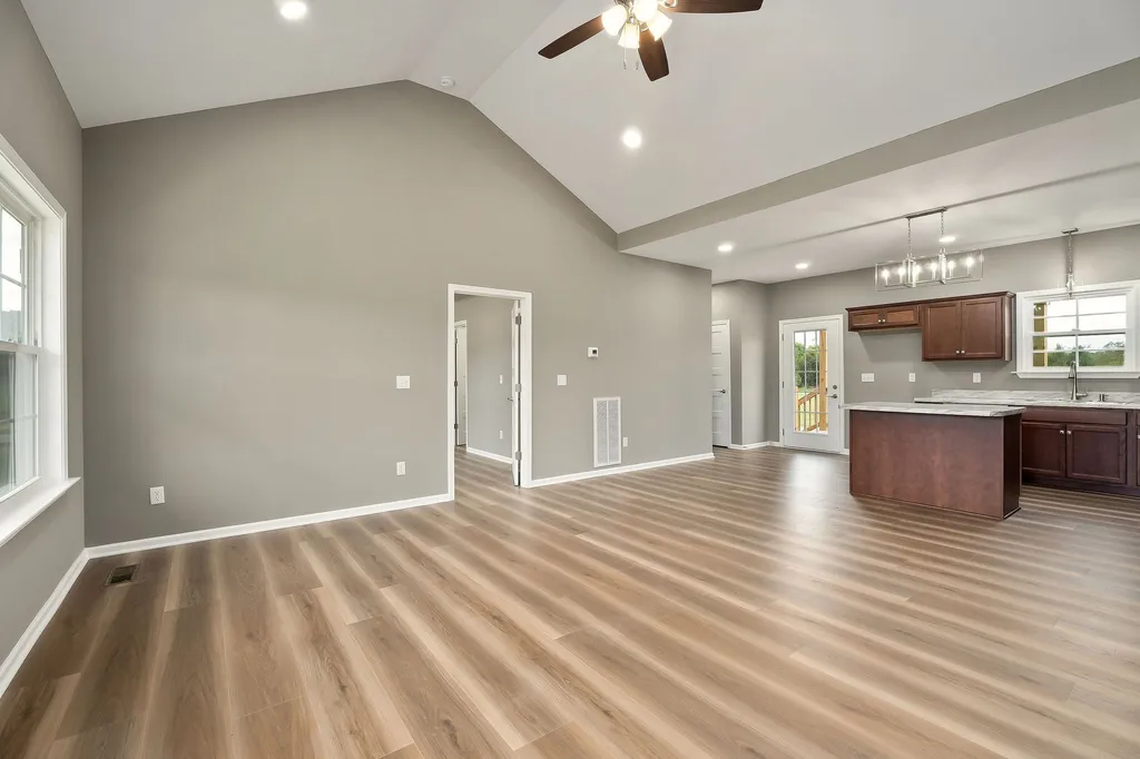 Spacious open-concept room with light wood flooring, gray walls, and a vaulted ceiling. A ceiling fan and recessed lighting create an airy feel. The kitchen area has dark wood cabinets, an island, and a chandelier. A door with glass panels leads outside, providing natural light.