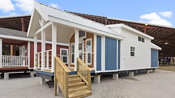 Cozy tiny house with blue and white siding, a small wooden porch, and stairs. The house is elevated on blocks, set under a clear blue sky.