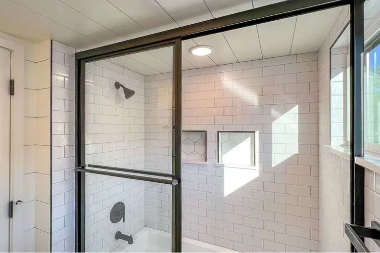 Modern shower with white subway tiles, black framed glass door, and built-in shelves. Soft sunlight casts shadows, creating a clean, airy feel.