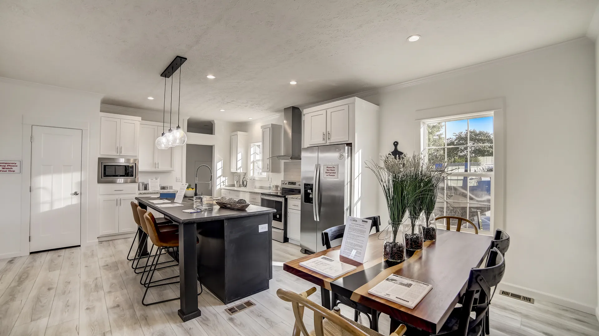 Modern kitchen with white cabinets, stainless steel appliances, and a central black island with pendant lights. Adjacent dining table with decorative plants. Bright, airy atmosphere.