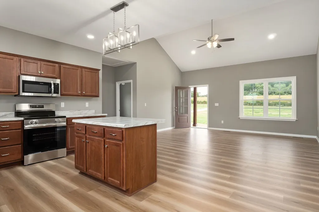 Modern open-concept living space with light wood floors. Features a kitchen with cherry cabinets, marble island, stainless steel appliances, and bright natural light from large windows.