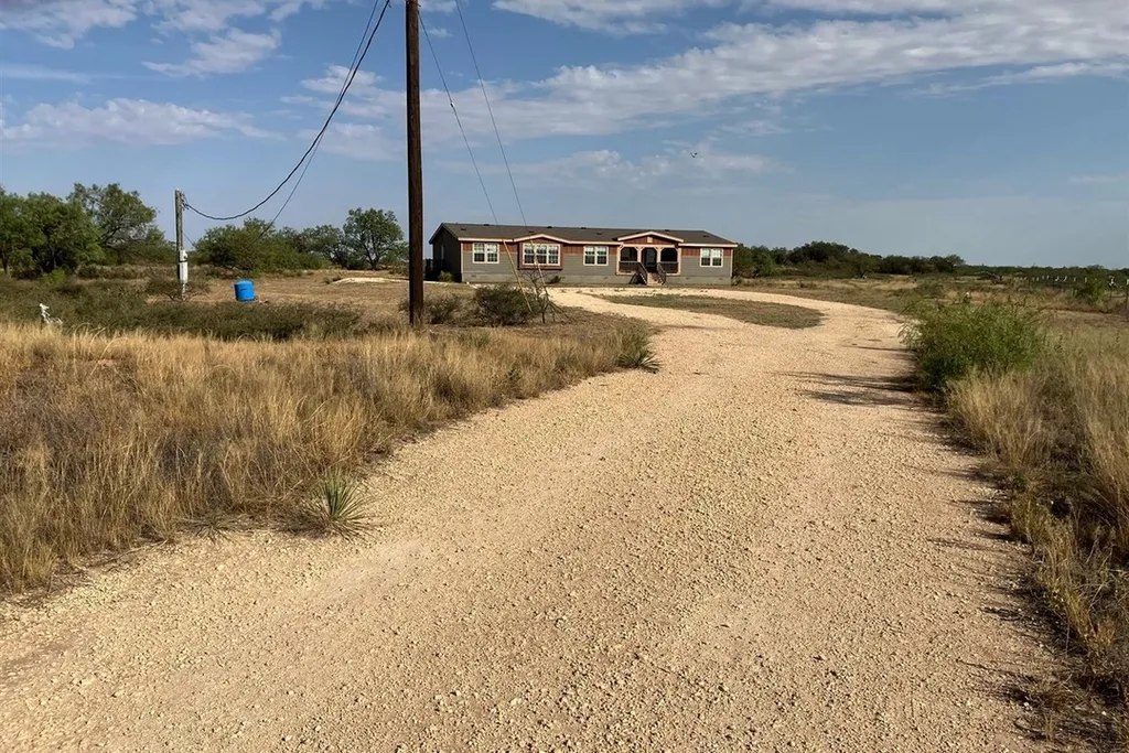 A long gravel driveway leads to a beige ranch-style house in a rural landscape. Dry grass and bushes border the path under a clear blue sky.