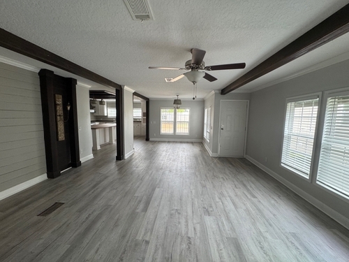 Spacious empty living room with light gray wood flooring, white walls, ceiling fan, and large windows with blinds, creating a bright, open atmosphere.