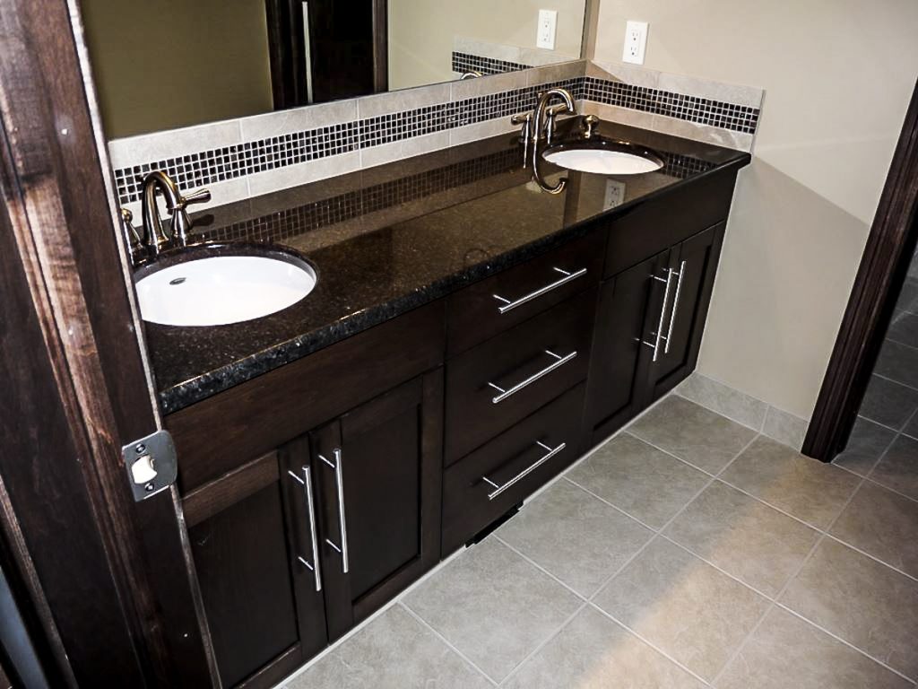Modern bathroom with a dark wood double vanity, black granite countertop, two sinks, silver faucets, mosaic tile backsplash, and grey tile floor.