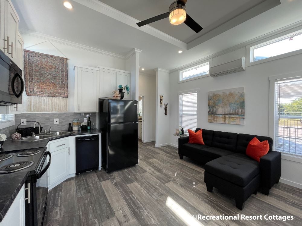 A modern kitchen and living area with white cabinets, black appliances, and gray wood floors. A black sectional sofa with orange pillows sits near large windows, creating an inviting and cozy atmosphere.