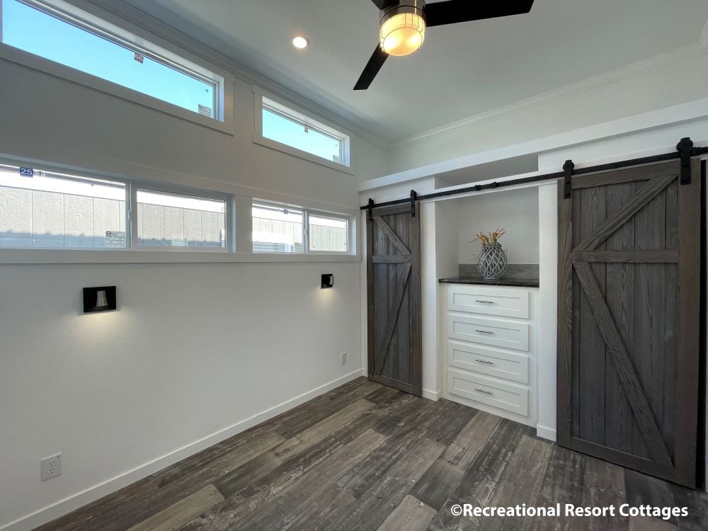 Modern, minimalist room with high windows, dark wooden sliding barn doors, white drawers, and gray wood floor. Ceiling fan adds a cozy touch.