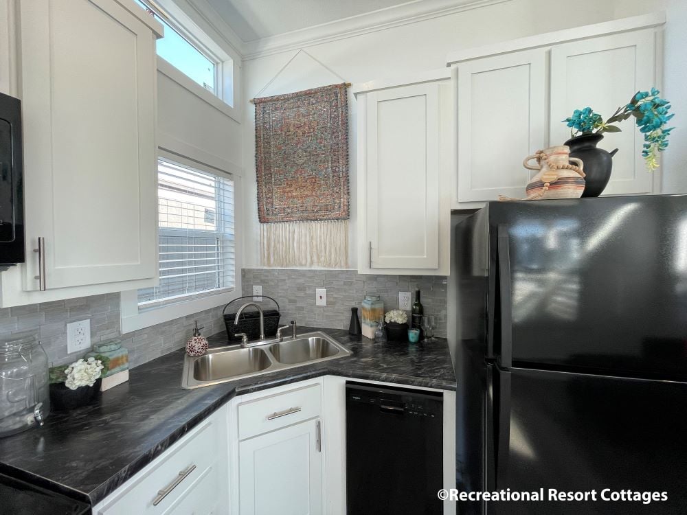 Compact kitchen with white cabinets and dark countertops. A black fridge and double sink are present. Wall art, plants, and pottery add warmth. Cozy atmosphere.