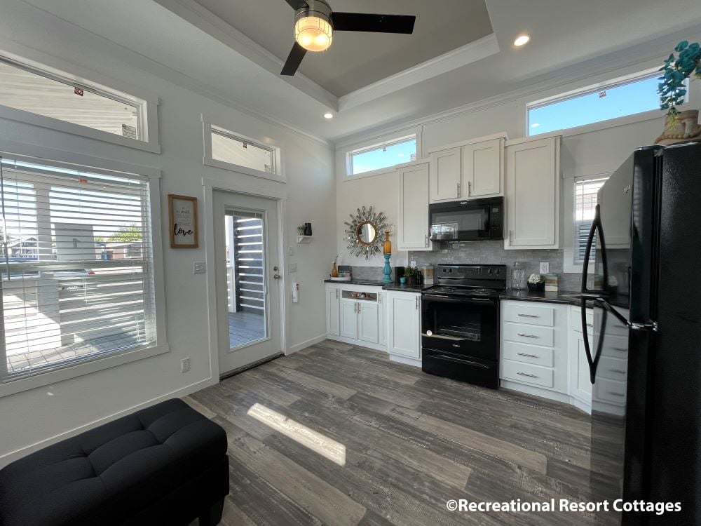 Modern kitchen with white cabinets, black appliances, and wood-look flooring. Sunlight streams through large windows, creating a bright, airy feel. A circular mirror and decor add charm.