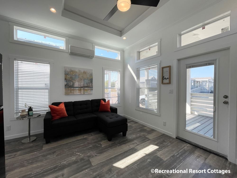 Modern living room with a black sectional sofa, red cushions, and a small side table. Natural light streams through large windows, creating a warm, inviting atmosphere.