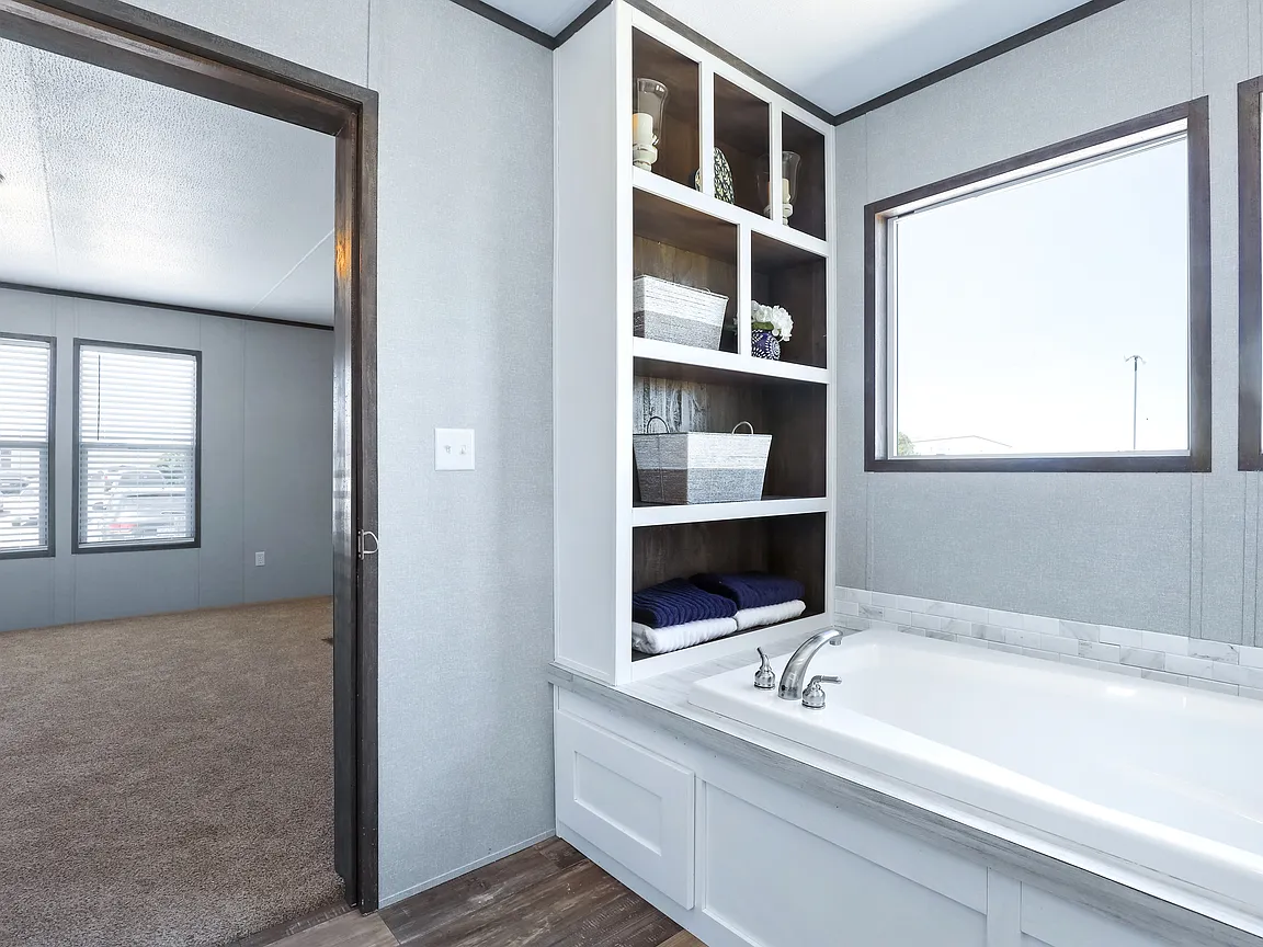 Modern bathroom view with a white bathtub, built-in shelving holding towels and baskets, and a large window. Adjacent room visible through an open door.